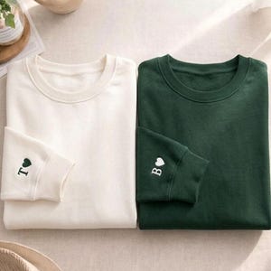 Op de afbeelding: Twee crewneck sweatshirts, één crème en één bosgroen, worden getoond. Het crèmekleurige sweatshirt heeft de letter "T" en een hartje geborduurd op de manchet. Het groene sweatshirt heeft de letter "B" en een hartje geborduurd op de manchet.