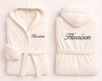 May include: A cream-coloured hooded bathrobe featuring a tie belt and long sleeves. The front left chest and the back of the robe are embroidered with the name Harrison in a dark grey cursive script font, showcasing both front and back views.