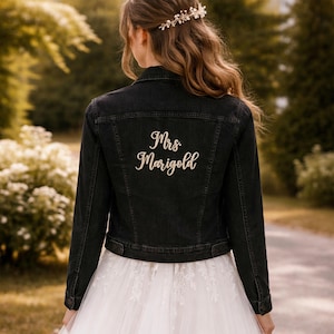 May include: A black denim jacket with the embroidered text "Mrs. Marigold" in a cursive font. The jacket is worn over a white wedding dress. The image is taken outdoors with a blurred background of trees and foliage.