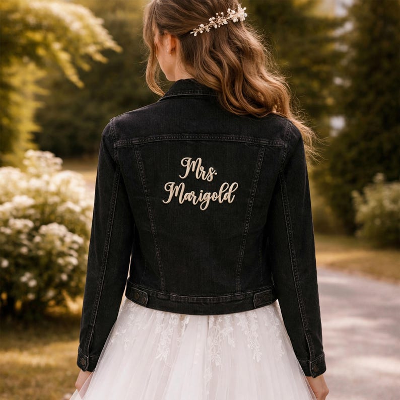 May include: A black denim jacket with the embroidered text "Mrs. Marigold" in a cursive font. The jacket is worn over a white wedding dress. The image is taken outdoors with a blurred background of trees and foliage.