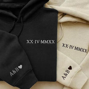 May include: Black and beige hoodies with white text. The black hoodie has "XX-IV-MMXX" on the chest and "A&B❤" on the sleeve. The beige hoodie has "XX-IV-MMXX" on the chest and "A&B❤" on the sleeve. The hoodies are made of a soft material.
