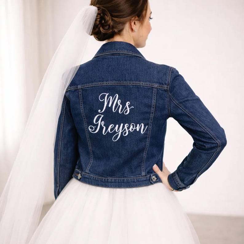 May include: A dark blue denim jacket with the embroidered text "Mrs Greyson" in white script. The jacket has a classic design with button closures and a collar. The bride is wearing a white tulle skirt and veil.