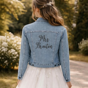 May include: Light blue denim jacket with the embroidered text "Mrs. Braxton" on the back. The jacket has a classic design with button closures and a collar. The jacket is worn over a white wedding dress.