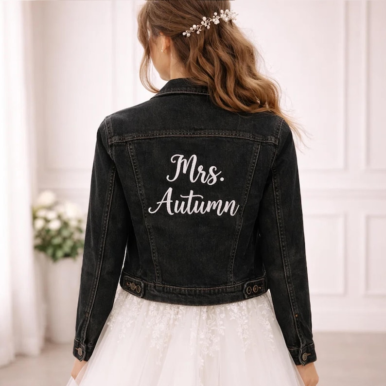 May include: A black denim jacket featuring the white text Mrs. Autumn printed on the back. The jacket has a classic collar, long sleeves with buttoned cuffs, and a buttoned waistband. It is worn over a white dress with lace details.