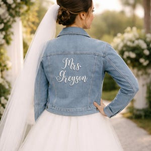 May include: A light blue denim jacket with the embroidered text "Mrs. Greyson" on the back. The jacket is worn over a white wedding dress. A white veil is draped over the shoulder.
