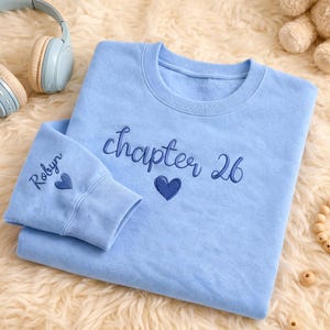 Puede incluir: Sudadera azul claro con la inscripción bordada "chapter 26" y un corazón. El puño de la manga lleva el nombre "Robyn" y un corazón. La sudadera es de un tejido suave.