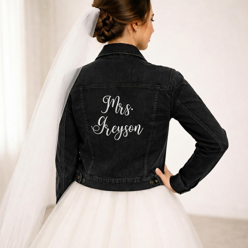May include: Black denim jacket with the text "Mrs. Greyson" in white script on the back. The jacket has a classic design with button closures and a collar. The bride is wearing a white tulle skirt and veil.