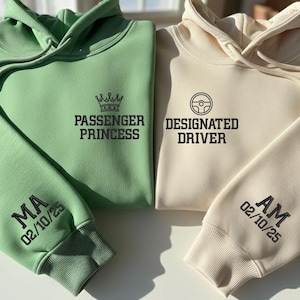 Può includere: Due felpe con cappuccio, una verde chiaro e una color crema. La felpa verde ha "PASSENGER PRINCESS" stampato sul davanti e "MA 02/10/25" sulla manica. La felpa color crema dice "DESIGNATED DRIVER" e "AM 02/10/25" sulla manica.
