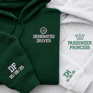 Könnte beinhalten: Zwei Kapuzenpullover, einer dunkelgrün und einer weiß. Der grüne Hoodie trägt die Aufschrift "DESIGNATED DRIVER" mit einem Lenkrad-Grafik. Der weiße Hoodie trägt die Aufschrift "PASSENGER PRINCESS" mit einer Kronen-Grafik. Beide Ärmel haben Initialen und ein Datum.
