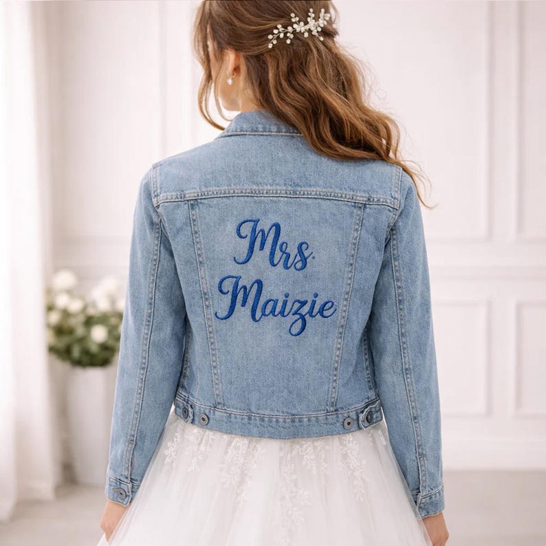 May include: Light blue denim jacket with long sleeves. The back of the jacket features the embroidered text "Mrs Maizie" in a vibrant blue. The jacket is buttoned and appears to be part of a bridal ensemble.