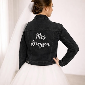 May include: A black denim jacket with the embroidered text "Mrs Greyson" in white script. The jacket has a classic design with button closures and pockets. The bride is wearing a white tulle wedding dress and veil.