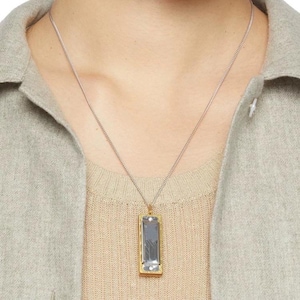 May include: A silver chain necklace with a rectangular pendant. The pendant has a gold-colored frame and a rectangular, light blue-gray stone. The necklace is worn over a beige sweater and a light gray jacket.