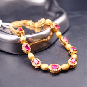Könnte beinhalten: Ein goldenes Armband mit ovalen, rubinroten Edelsteinen, umgeben von kleinen weißen Steinen. Das Armband hat eine feine Kette und liegt auf einer dunklen Oberfläche. Das Armband ist ein Schmuckstück.