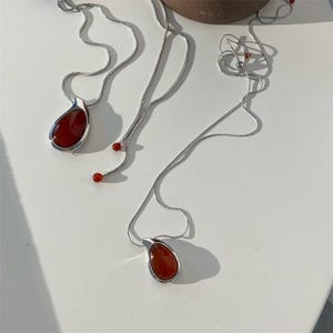 May include: Three silver necklaces with teardrop-shaped, red gemstone pendants. One necklace has two small red beads. The necklaces are displayed on a white surface. The pendants are approximately 1 inch in length.
