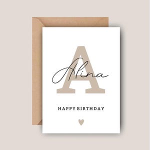 Könnte beinhalten: Eine weiße Grußkarte mit einem beigefarbenen Umschlag. Die Karte zeigt einen großen, beigefarbenen Buchstaben "A" mit dem Namen "Alina" in Schreibschrift. Darunter stehen die Worte "HAPPY BIRTHDAY" und ein kleines Herz.