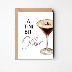 Könnte beinhalten: Grußkarte mit einem braunen Kraftpapierumschlag. Die Karte zeigt eine Aquarellillustration eines Martinis und den Text "A Tini Bit Older" in Schwarz. Der Martini hat eine dunkelbraune Flüssigkeit und drei Kaffeebohnen.