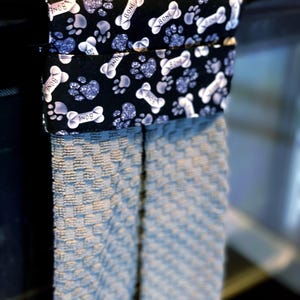 May include: A kitchen towel holder with a black and white dog bone and paw print pattern. The holder is attached to a light blue patterned kitchen towel. The words "BONE" are printed on the dog bones.
