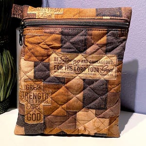 May include: A quilted pouch in shades of brown, tan, and black, featuring a patchwork design. The pouch has a zipper closure and includes text that reads "Be Strong and Courageous" and "Great Strength Comes From Faith in God."