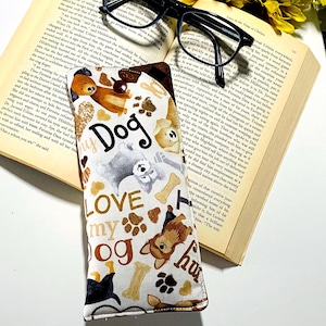 May include: A fabric glasses case with a white background, featuring a pattern of brown and gray dogs, paw prints, and bones. The words "LOVE my DOG" are printed on the case. Black eyeglasses rest on an open book.