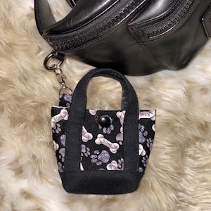 May include: A small black tote bag with a black handle and a button closure. The bag features a pattern of white bones, paw prints, and silver glitter on a black background. The bag is attached to a black leather fanny pack.