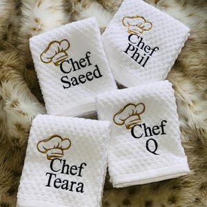 May include: Four white kitchen towels with a waffle weave texture. Each towel features an embroidered chef's hat and the name "Chef" followed by a name: Saeed, Phil, Teara, and Q. The embroidery is in gold and black.
