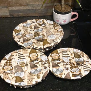 May include: Three round, beige fabric covers with a brown and white gnome pattern. The gnomes are holding coffee cups and bags of coffee. The fabric covers have a ruffled edge.