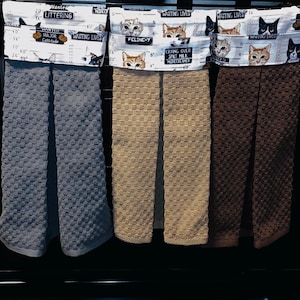 May include: Three hanging kitchen towels in gray, tan, and brown with a waffle-textured weave. Each towel features a white fabric topper printed with cat mugshots, height charts, and text including Wanted, Major Cattitude, Feline-Y, and Wasting Lives.