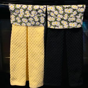 May include: Two kitchen towels hanging on a rack. The towel on the left is yellow with a white daisy print. The towel on the right is black with a white daisy print.