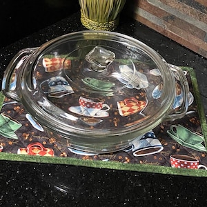 May include: A clear glass casserole dish with a matching lid featuring two side handles, resting on a rectangular fabric mat. The mat has a dark brown background with a repeating pattern of colorful coffee cups, saucers, and scattered coffee beans.