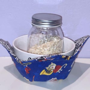 May include: A white ceramic bowl with a blue and white fabric bowl cozy. The cozy has a pattern of cats playing musical instruments. A glass jar with a silver lid is inside the bowl, filled with oats.