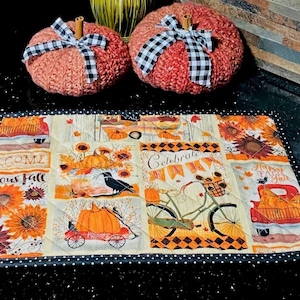 May include: Two crocheted orange pumpkins with cinnamon stick stems and black and white buffalo check bows, placed above a rectangular quilted fabric mat featuring sunflowers, pumpkins, a bicycle, a red truck, and the text Celebrate Fall.