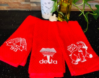 May include: Three red kitchen towels with white embroidered designs. The first towel has two hands forming a triangle with the year 1913 below. The second towel has a book with the word "delta" below. The third towel has an elephant with the year 1913 above.