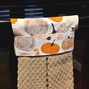 May include: A hanging kitchen towel featuring a top fabric panel with a white background and a pattern of orange and tan pumpkins with brown stems. The lower section consists of a textured, tan-colored waffle-weave fabric towel.