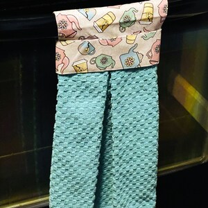 May include: A teal kitchen towel with a patterned fabric band featuring a teacup and teapot design. The towel is hanging on a black oven door.