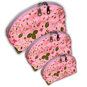 May include: Three pink cosmetic bags with a floral strawberry pattern. The bags are different sizes and have silver zips. The fabric features green leaves, white flowers, and red strawberries.