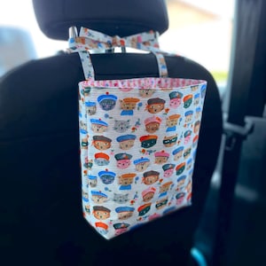 May include: A car seat organizer with a whimsical cat print. The fabric features various cat faces wearing hats and berets on a white background with blue stripes. The organizer hangs from the headrest with a matching patterned bow.