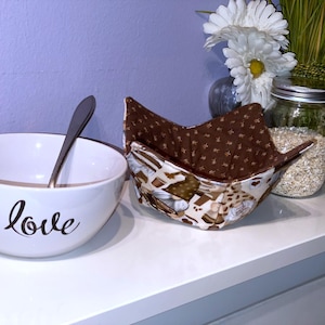 May include: A white ceramic bowl with the word "love" in cursive script, a silver spoon, and two fabric bowl cozies. The cozies have a brown and cream pattern. A jar of oats and flowers are in the background.