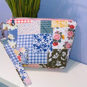 May include: A patchwork cosmetic bag with a wrist strap. The bag features a variety of floral and gingham patterns in blue, pink, white, and yellow. A pink zipper and silver hardware complete the design. The bag is sitting on a white surface.