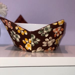 May include: A brown fabric bowl cozy with a white interior. The exterior features a pattern of paw prints in yellow, orange, and green, set against a dark brown background. The cozy is designed to hold a bowl and keep food warm.