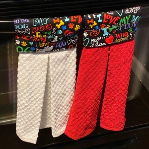 May include: Two kitchen towels hanging on a black oven handle. The towel on the left has a black background with colorful text that says "I love my dog" and paw prints. The towel on the right has a red background with colorful text that says "I love my dog" and paw prints.