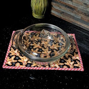 May include: A clear glass casserole dish with a lid resting on a rectangular fabric mat. The mat features a black background with a repeating pattern of gingerbread people and white snowflakes, finished with a red and white striped fabric border.