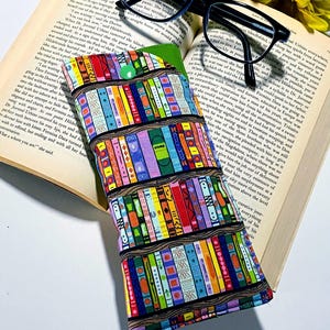 May include: A colorful glasses case with a bookcase design rests on an open book, next to black-framed glasses and yellow flowers. The case features a green interior and a button closure, offering a stylish way to protect eyewear.