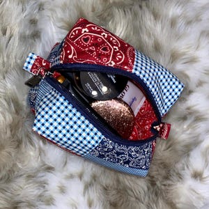 May include: A blue and red patchwork zippered pouch with a bandana print. The pouch is open and contains various makeup products, including a compact, a bottle of liquid, and a jar of glitter.
