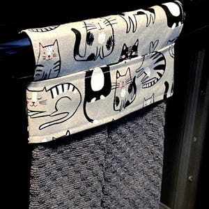 May include: A hanging kitchen towel featuring a light gray fabric top with a repeating pattern of black, white, and gray cartoon cats. The towel has a textured, dark gray absorbent bottom section and is draped over a black horizontal bar.