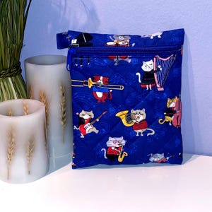 May include: A blue quilted pouch with a zipper closure. The pouch is decorated with a pattern of cats playing musical instruments. The cats are dressed in various outfits and are playing instruments such as a guitar, harp, and saxophone.