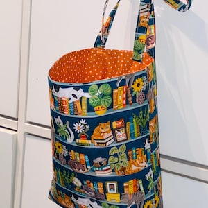 May include: A fabric tote bag with a teal background featuring a bookshelf design with cats, books, and plants. The interior is orange with white polka dots. The straps and top trim have a coordinating floral pattern. The bag is hanging from a decorative knob.