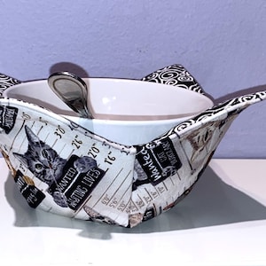 May include: A fabric bowl cozy featuring a white background with a repeating print of cats in mugshot poses, including text that reads "WANTED" and "WASTING LIVES." The cozy has black and white swirl patterned corners and holds a white ceramic bowl.