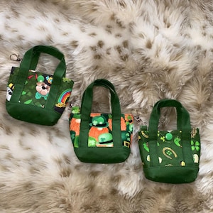 May include: Three small green tote bags with matching handles. Each bag features a different St. Patrick's Day themed pattern, including shamrocks, leprechauns, and rainbows. The bags are made of fabric and have a rectangular shape.