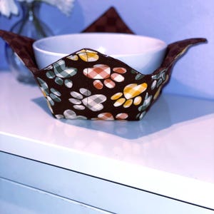 May include: A white ceramic bowl sits inside a brown fabric bowl cozy. The cozy is patterned with colorful paw prints in white, yellow, orange, and green. The cozy has extended flaps on each side. A brown triangle is behind the bowl.