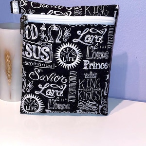 May include: A small, rectangular black pouch with a white zipper and a white pull tab. The pouch is covered in white script and illustrations, including the words "Jesus", "Lord", and "King". The design has a religious theme.